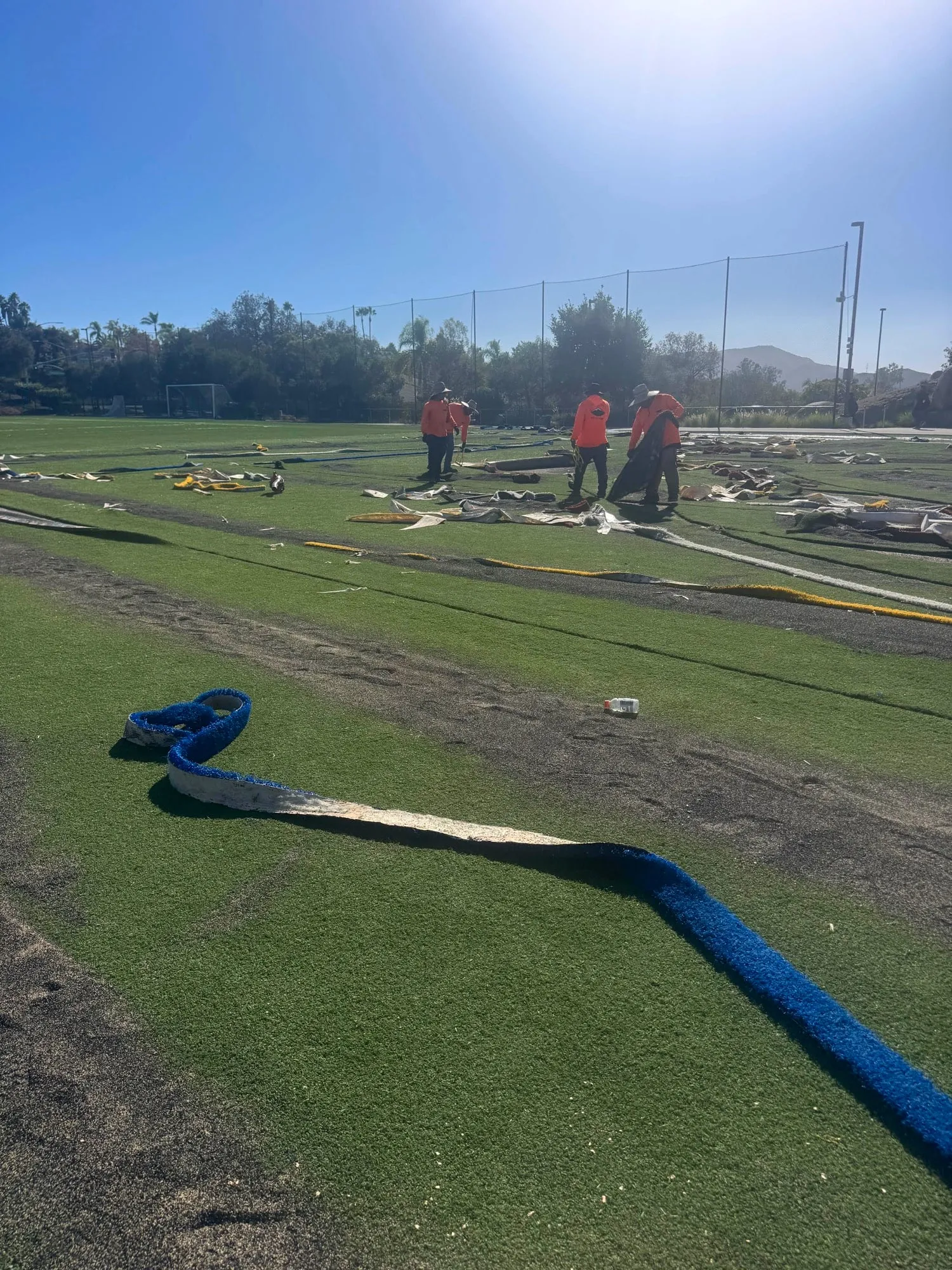 Crew in orange shirts pulling old seam tape and inlay strips off a synthetic field